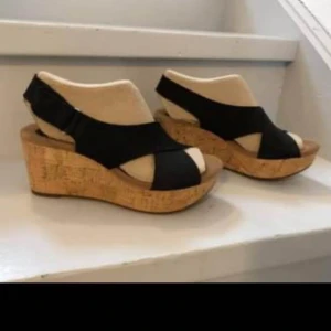 Svarta sandaler i skinn med kilklack från Clarks strl 39 -  Snygga svarta sandaler från Clarks helt i skinn med bekväm kilklack i kork. De har en korsad design över foten och en justerbar rem runt hälen. Perfekta för sommarens alla tillfällen.Aldrig använda, pga för små.