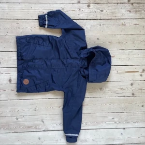 Blå vindjacka från Mini Rodini - Säljer en blå vindjacka från Mini Rodini med avtagbar huva och reflexdetaljer på ärmarna. Jackan har en praktisk dragkedja och knappar framtill. Perfekt för blåsiga dagar! 💙