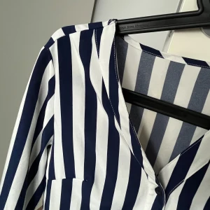 Randig blus i blått och vitt - Snygg blus med vertikala ränder i blått och vitt. Blusen har långa ärmar och en v-ringad design framtill. Perfekt för en stilren look.