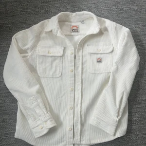 Vit manchester overshirt från Deus - Säljer en stilren vit manchester overshirt från Deus. Skjortan har långa ärmar och två bröstfickor med knappar. Perfekt som overshirt aldrig använd pga för stor för mig
