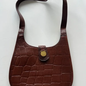 Brun axelväska i skinn från Mulberry - Snygg brun axelväska i skinn från Mulberry med krokodilmönster. Väskan har ett elegant guldspänne och ett rutigt innerfoder med dragkedja. Perfekt för att ge en stilren touch till din outfit.