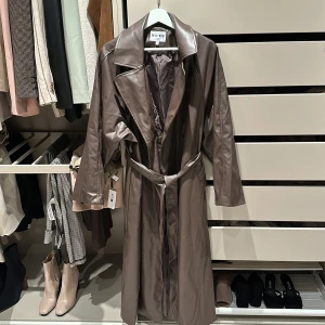 Brun trenchcoat från NA-KD - Snygg brun trenchcoat från NA-KD i syntetmaterial. Jackan har långa ärmar och ett bälte i midjan för en stilren look. Perfekt för höst och vår.