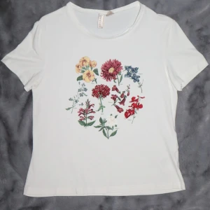 Vit t-shirt med blommigt tryck - Vit t-shirt med ett färgglatt blommigt tryck på framsidan. T-shirten är i bomull och har en normal passform med korta ärmar. Den är i bra skick!