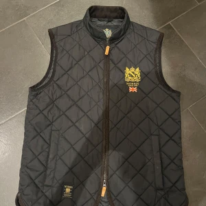  quiltad väst från Morris - Säljer en snygg svart quiltad väst från Morris i storlek medium. Västen har en dragkedja framtill och ett stilrent emblem på bröstet. Perfekt för höst och vår när du vill hålla dig varm men ändå se stilren ut. Passar både till vardags och lite finare tillfällen. Köpt för 1500 ish. 