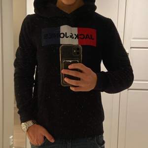 Säljer en svart hoodie från Jack & Jones med en cool logga i rött, vitt och blått på bröstet. Den är perfekt för både höst och vinter men funkar när som helst. Tröjan har en skön passform med långa ärmar och en praktisk ficka framtill. Perfekt för en avslappnad stil!