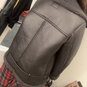 Svart skinnjacka med foder - Säljer en snygg svart skinnjacka med mjukt foder och bälte i midjan. Jackan har en cool, oversized look med långa ärmar och dragkedja framtill. Perfekt för höst och vinter när du vill hålla dig varm och stilren. Passar både till vardags och fest!