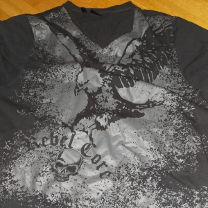 Svart t-shirt med örntryck - Cool svart t-shirt från Rebel Core med ett stort tryck av en örn och en dödskalle. Den har en v-ringad hals och är perfekt för en avslappnad stil. Passar bra till jeans eller shorts för en rockig look.