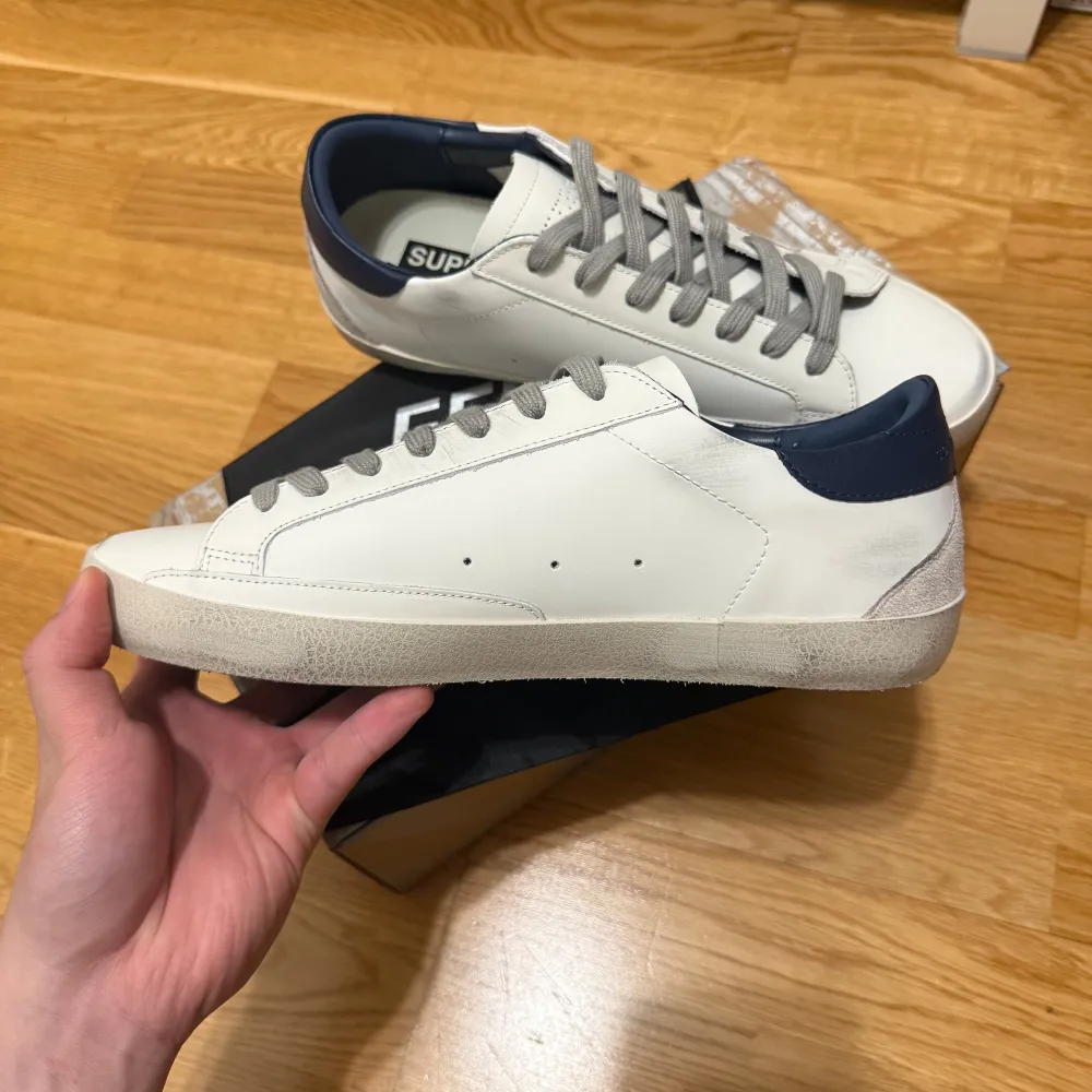 Säljer ett par vita Golden Goose sneakers i mycket bra skick. De har en snygg stjärndetalj på sidan och grå snörning. Hälpartiet är i mörkblått skinn och sulan har en lätt sliten look för den där coola vintagekänslan. Perfekta för både vardag och fest! Storlek 42. Skor.