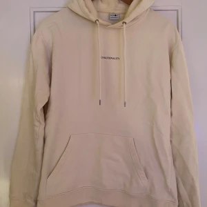Beige hoodie från NN07 - Säljer en stilren beige hoodie från NN07 i mycket bra skick. Den har en klassisk design med känguruficka och justerbar huva med dragsko. Perfekt för både vardag och mysiga stunder. Texten 'No Nationality' pryder bröstet för en minimalistisk look.