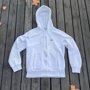 Vit hoodie från Lacoste - Säljer en klassisk vit hoodie från Lacoste i storlek M. Den har en dragkedja framtill och en praktisk känguruficka. Perfekt för höst och vår! Lite använd men i bra skick. 🐊