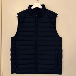 Marinblå quiltad dunväst - Säljer en stilren marinblå quiltad dunväst i mycket bra skick. Västen har en dragkedja framtill och är perfekt för höst och vår. Västen är ifrån Kappahl och har aldrig använts 