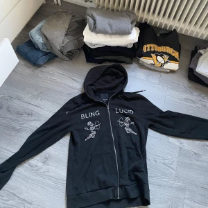 Svart rhinestone hoodie från Bling Lucid - Säljer en svart rhinestone hoodie från Bling Lucid i storlek M. Den har en dragkedja framtill och coola detaljer med texten 'BLING' och 'LUCID' på bröstet. Använd få gånger så i bra skick 