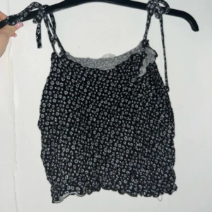Svart blommig topp från & Other Stories - Säljer en supersöt svart topp med vita blommor från & Other Stories. Den har smala axelband som knyts och en elastisk, smockad design för en bekväm passform. Perfekt för sommardagar eller en avslappnad kväll ute. 🌼
