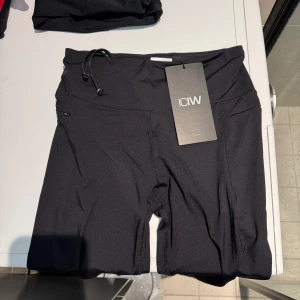 Svarta träningstights från ICANIWILL - Säljer ett par svarta träningstights från ICANIWILL i nytt skick. De är högmidjade med en praktisk dragsko i midjan för bästa passform. Materialet är stretchigt och perfekt för träning. Shortsen har en diskret ficka på sidan för småsaker samt en dragkedja på ankeln. Perfekta för gymmet eller löprundan!