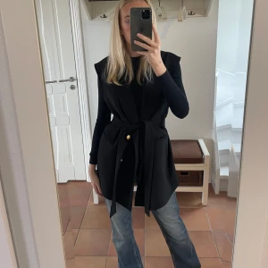 Svart väst med knytband - Säljer en stilren svart väst med knytband i midjan. Perfekt för lager-på-lager looken och passar både till vardags och fest. 