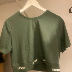 Grön kortärmad topp från Stronger - Säljer en grön kortärmad topp från Stronger i storlek L. Toppen är tillverkad av 90% återvunnen polyester och 10% elastan, vilket gör den både bekväm och stretchig. Perfekt för träning eller en avslappnad dag. Den har texten 'STRONGER' tryckt längs fållen.
