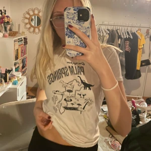 Vit t-shirt med Palm Springs-tryck - Säljer en vit t-shirt med coolt Palm Springs-tryck. Den har en avslappnad passform och är perfekt för sommardagar. Trycket har en retro känsla med palmer och dinosaurier. Superbekväm och lätt att matcha med jeans eller shorts!