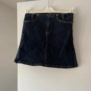 Mörkblå jeanskjol - Säljer en snygg mörkblå jeanskjol i mycket bra skick. Kjolen har en klassisk design med fem fickor och en liten slits på sidan för extra rörelsefrihet. Perfekt för både vardag och festliga tillfällen. Passar bra till en t-shirt eller en stickad tröja för en mer avslappnad look.
