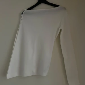 Vit oneshoulder topp med knappar - Säljer en stilren vit oneshoulder topp med knappar. Toppen är i ett mjukt material och passar både till vardags och fest. Helt ny!