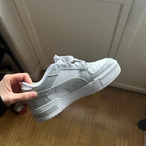 Vita sneakers från Puma - Säljer ett par snygga vita sneakers från Puma i mycket bra skick. De har en klassisk design med snörning och en bekväm sula. Perfekta för vardagsbruk och passar till nästan alla outfits. Storleken är inte angiven, men de ser ut att vara i standardstorlek. Skorna är i syntetmaterial och har en stilren look.
