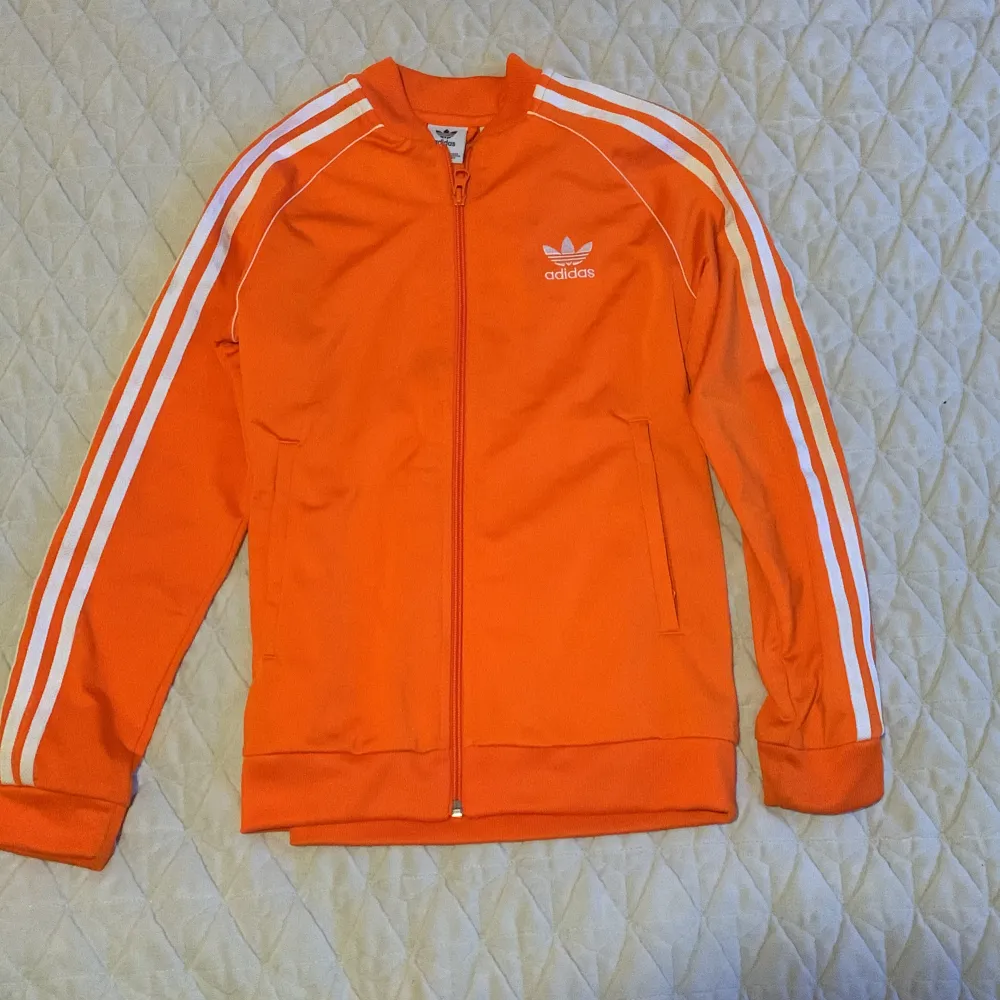 Säljer en snygg orange träningsjacka från Adidas med de klassiska tre ränderna längs ärmarna. Jackan har dragkedja och är perfekt för sportiga aktiviteter. Den är i mycket bra skick och passar perfekt för höst och vår.. Sportswear.