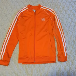 Orange träningsjacka från Adidas storlek 152 cm - Säljer en snygg orange träningsjacka från Adidas med de klassiska tre ränderna längs ärmarna. Jackan har dragkedja och är perfekt för sportiga aktiviteter. Den är i mycket bra skick och passar perfekt för höst och vår.