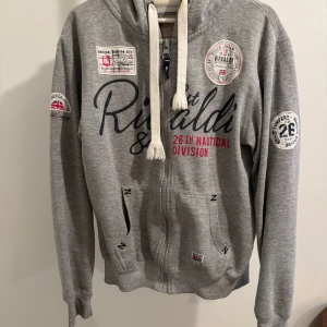 Grå hoodie - Säljer en snygg grå hoodie med dragkedja och stora tryck på både fram- och baksidan. Den har en cool marin stil med text och nummerdetaljer. Perfekt för en avslappnad look!