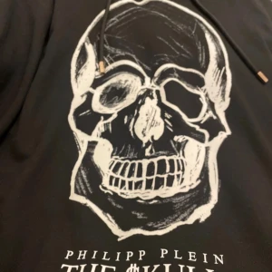 Svart hoodie från Philipp Plein med dödskallemotiv - Säljer en cool svart hoodie från Philipp Plein med ett stort dödskalletryck på framsidan. Den är i 100% bomull och har en skön passform med långa ärmar och justerbar huva. Perfekt för en avslappnad stil eller en utekväll. Hör av dig vid intresse!