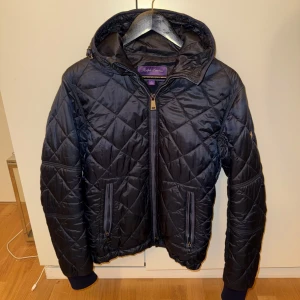 Marinblå quiltad jacka från Ralph Lauren Purple Label - Säljer en snygg svart quiltad jacka från Ralph Lauren i storlek S. Jackan har en dragkedja framtill och en mysig huva. Perfekt för höst och vår, med en stilren look som passar till det mesta. Den är i mycket bra skick och redo för nya äventyr!