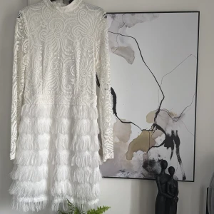 Vit spetsklänning - Säljer en superfin vit klänning med långärmad design och vackra spetsdetaljer. Klänningen har en unik fransad nederdel som ger en härlig rörelse. Perfekt för festliga tillfällen eller en elegant sommarkväll. Den är i mycket bra skick och redo att bäras av någon som vill sticka ut!, (ordinarie pris 550)