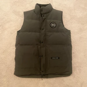 Canada Goose black label väst  - Säljer en snygg grå dunväst från Canada Goose i mycket bra skick. Västen har en broderad logga på bröstet och praktiska fickor framtill. Perfekt för kyliga höst- och vinterdagar. Den är i storlek M. Dragkedjan är lite trög men annars är allt perfekt med den. Priset är diskuterbart vid snabb affär. 