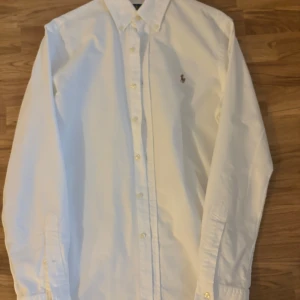 Vit skjorta från Ralph Lauren - Säljer en klassisk vit skjorta från Ralph Lauren i slim fit. Skjortan är långärmad och har knappar hela vägen fram. Den är i bomull och har den ikoniska broderade loggan på bröstet. Perfekt för både vardag och fest! 🕺