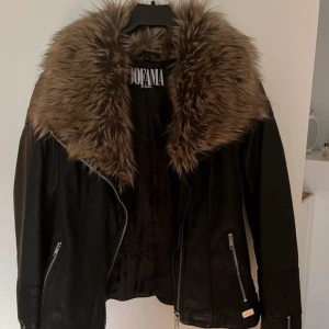 Svart skinnjacka från Jofama by Kenza  - Säljer en snygg svart skinnjacka från Jofama by Kenza. Jackan har en stor, fluffig päls i brunt runt kragen och dragkedja framtill. Den är i mycket bra skick då jag endast har använt den en gång sedan har den bara hängt i garderoben och nu är den för liten för mig. Den är perfekt för höst och vinter. Passar både till vardags och fest!