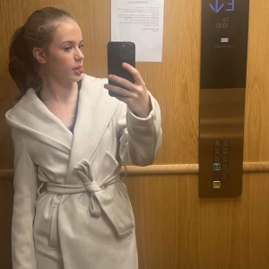 Crémevit kappa från Zara - Säljer en superfin vit kappa från Zara i storlek XS. Den har en snygg omlott-design med ett bälte i midjan och en stor huva. Perfekt för höst och vår när du vill vara både stilren och varm. Kappan är i mycket bra skick och gjord av ett mjukt material som känns lyxigt. Passar perfekt till både vardag och fest! Den har inga defekter och är i princip helt ny. Köpte den för mindre än ett år sen och säljer den pga att jag har köpt en ny jacka och inte behöver denna. 