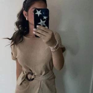 Beige bluse med bälte  - Säljer en stilren beige klänning med korta ärmar och ett snyggt bälte i midjan. Perfekt för både vardag och festliga tillfällen. Klänningen har en enkel och elegant design som passar till alla säsonger.