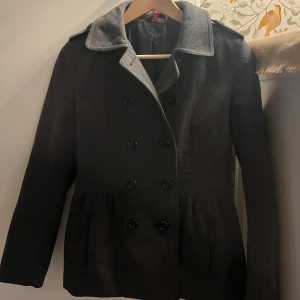 Grå figursydd kappa med knappar - Säljer en stilren svart kappa i mycket bra skick. Den har en klassisk design med dubbelknäppning och en fin krage. Fickor på sidorna. Perfekt för höst, vinter och vår, och passar både till vardags och fest. Storleken är dam S och materialet känns som ullblandning. Två extra knappar medföljer. Jag tror att den är från H&M. Säljer då jag inte använder den så mycket och behöver göra plats för annat.