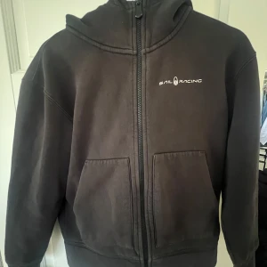 Svart hoodie från Sail Racing - Säljer en svart hoodie från Sail Racing i bra skick. Den har en dragkedja framtill och en praktisk huva. Perfekt för höst och vår, med en stilren design och logga på bröstet och huvan. Grym för både chill och aktivitet!