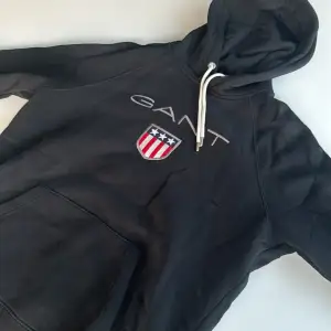 Säljer en snygg svart hoodie från GANT med ett coolt emblem på bröstet. Den har en stor ficka fram och justerbar huva med vita snören. Perfekt för en avslappnad stil eller kyliga dagar. 🖤