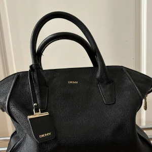 Svart handväska från DKNY - Snygg svart handväska från DKNY i skinn. Den har två handtag och en elegant design med subtila gulddetaljer. Insidan är fodrad med ett mönster av DKNY-loggan och har en praktisk dragkedja. Perfekt för både vardag och fest!