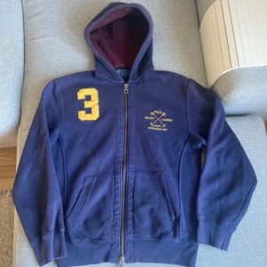 Mörkblå hoodie från Ralph Lauren - Säljer en snygg mörkblå hoodie från Ralph Lauren med dragkedja och gul detalj på bröstet. Den har en stor gul siffra 3 på ena sidan och en klassisk logga på andra. Perfekt för höst och vår, med en mysig luva i vinrött foder. Passar både till vardags och chill.