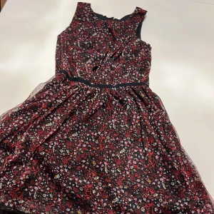 Blommig klänning från Name It - Säljer en superfin blommig klänning från Name It i storlek 11 år. Klänningen har en svart bas med ett färgglatt blommönster i rött, rosa och gult. Den är ärmlös och har en söt volangdetalj i midjan. Perfekt för vår och sommar! 🌸