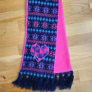 Desigual scarf - This is a stylish and colorful scarf with a unique design. The scarf has a pink fleece lining and a heart-shaped patch with the brand's name on one side. The other side has a circular patch with a similar design. The scarf has black tassels on both ends. Length: 105 cm. Wide: 17 cm.