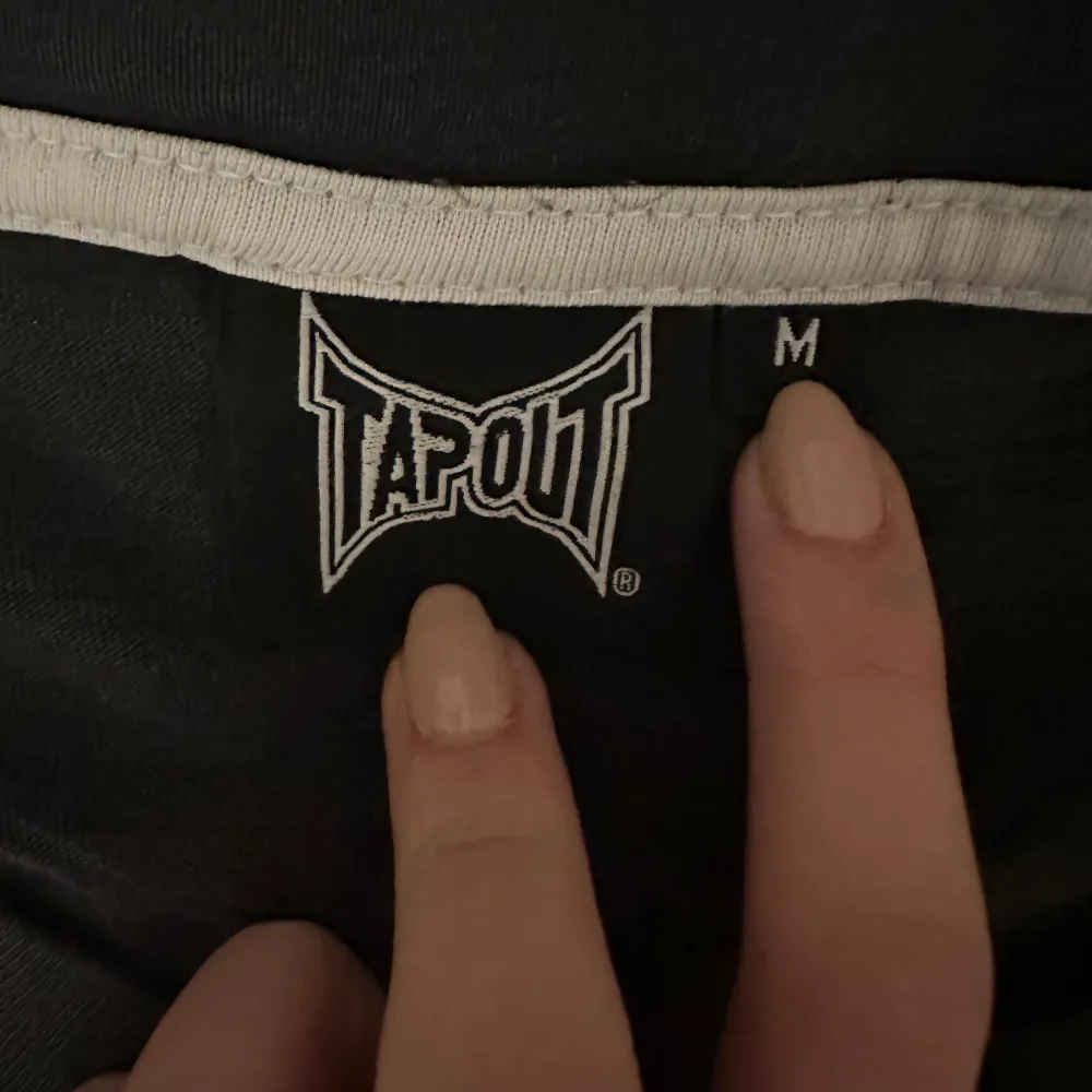 Säljer en svart T-shirt från Tapout i mycket bra skick. Den har ett stort, vitt Tapout-tryck på framsidan och ett mindre på baksidan. Perfekt för träning eller vardagsbruk. Materialet känns mjukt och bekvämt. Passar dig som gillar en sportig stil!. T-shirts.