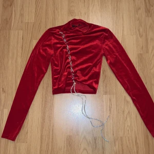 Röd sammetstopp från Soya Fashion - Snygg röd sammetstopp från Soya Fashion i storlek XS. Toppen har en unik snörning framtill med glittriga detaljer och långa ärmar. Perfekt för festliga tillfällen eller en utekväll. Den är både bekväm och stilren, och ger en lyxig känsla.
