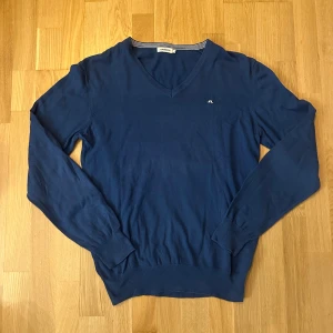 J.Lindeberg tröja  - En blå V-ringad tröja med J.Lindeberg logotypen på bröstet. Tröjan är lätt nopprig men syns inte så mycket. Se bild 4. Storlek M