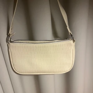 Beige axelväska med krokodilmönster - Snygg beige/vit axelväska i syntetmaterial med krokodilmönster. Väskan har en dragkedja upptill och en justerbar axelrem. Mellanstor väska, säljs då den inte kommer till användning längre.