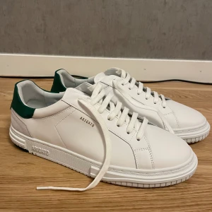 Vita sneakers från Axel Arigato - Säljer ett par stilrena vita sneakers från Axel Arigato i nytt skick, aldrig använda. De har gröna detaljer på hälen och är gjorda i skinn med snygg snörning. Perfekta för både vardag och fest! Storlek 46 men funkar med 45 om man har extra sula.