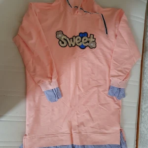 Rosa hoodie med paljettdetaljer - Säljer en superfin rosa hoodie med texten 'Sweet' i paljetter på framsidan. Den har långa ärmar och en cool detalj med randiga muddar i blått och vitt. Huvan har snören med texten 'LOVE'. Perfekt för en mysig dag eller en avslappnad utflykt! 💖