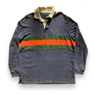 Blå pikétröja från Polo Ralph Lauren - Säljer en snygg blå pikétröja från Polo Ralph Lauren med en grön och röd rand över bröstet. Tröjan har en klassisk krage i beige och långa ärmar. Perfekt för en avslappnad men stilren look. Passar både till vardags och lite finare tillfällen. Storlek L.