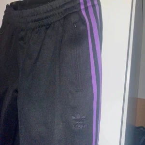 Svarta träningsbyxor från Adidas med lila ränder - Säljer ett par svarta träningsbyxor från Adidas i mycket bra skick. De har de klassiska tre lila ränderna längs sidorna och en elastisk midja för extra komfort. Perfekta för träning eller en avslappnad dag. Adidas-loggan är diskret placerad på benet.